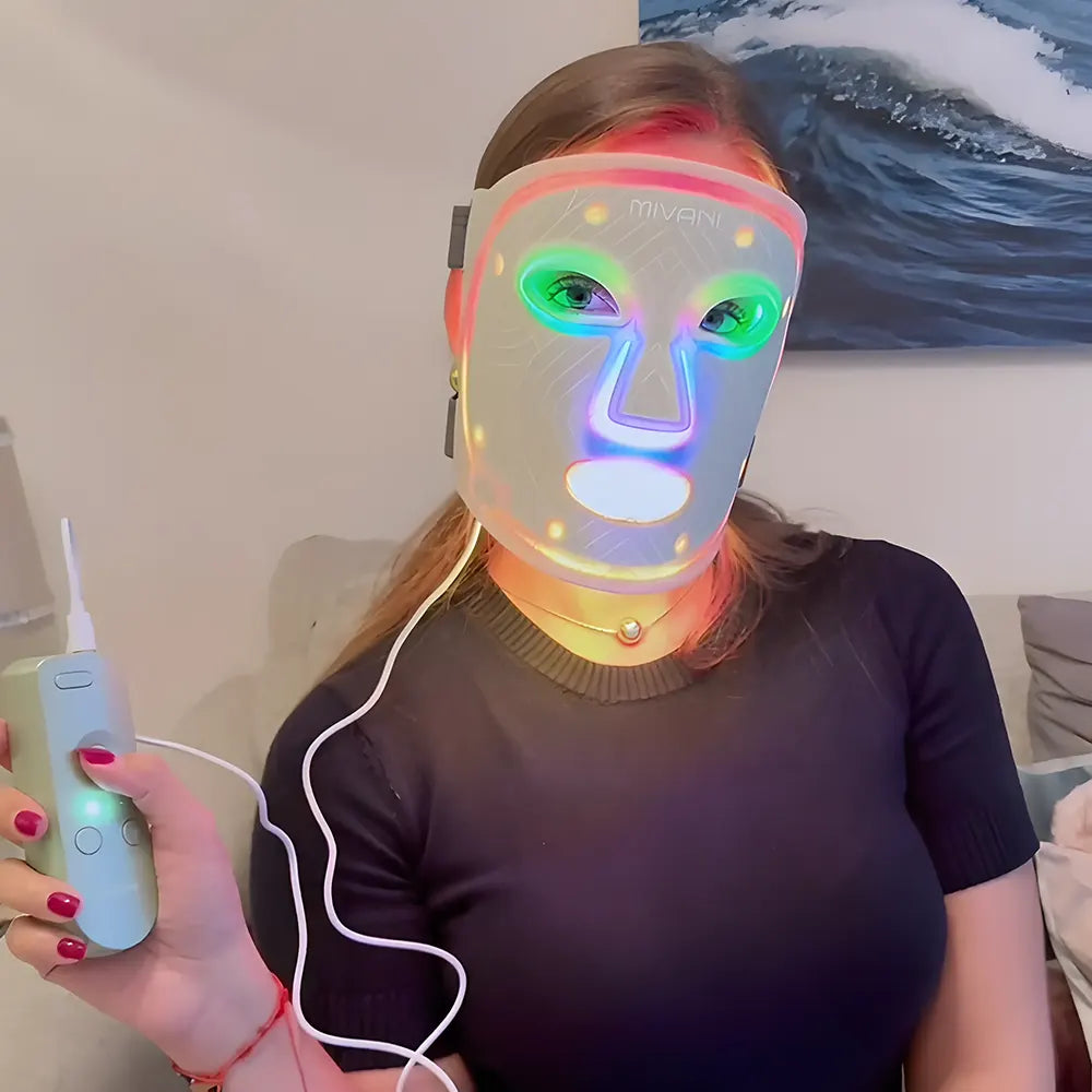 Person wearing a colorful LED face mask holding a remote control indoors.