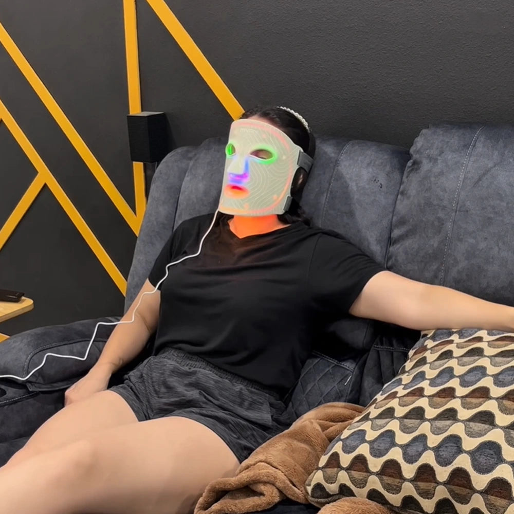 Woman sitting on a couch wearing a black shirt and gray shorts, with a colorful light therapy face mask on her face.