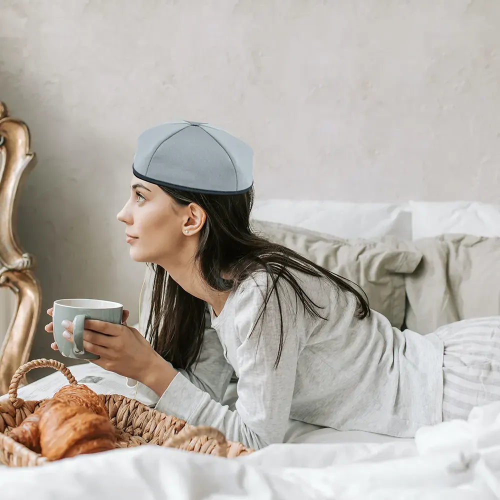 Woman in bed wearing a red and infrared light therapy hat and holding a mug, with a basket of bread on the side.