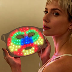 Woman holding a colorful LED mask against a plain background