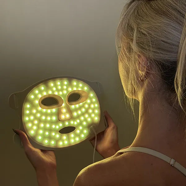 Person holding an amber glowing LED therapy face mask against a neutral background