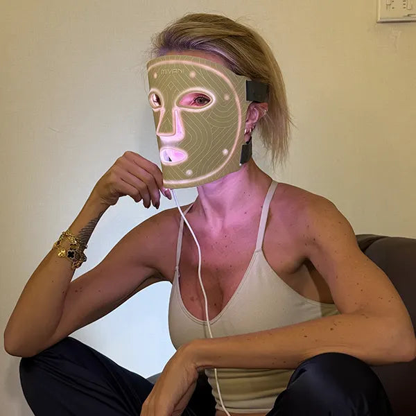 Person wearing a white LED therapy face mask with visible wires, sitting against a plain wall.