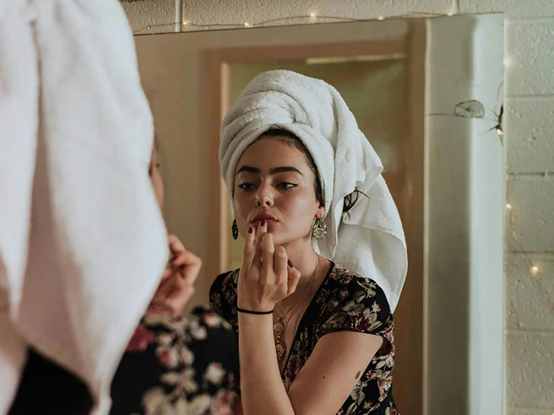 Woman applying makeup in front of a mirror with a towel on her head.
